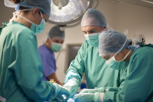 a female operating nurse stands over a patient at the operating table and looks down to what she is doing in the operation. She is joined by a young female nurse and mature male surgeon .In the background the anaesthetist is looking over from his monitors . They are all wearing surgical gowns and protective masks and headwear. they are wearing green gowns . The shot is horizontal waist up , with defocussed background for copy space .
